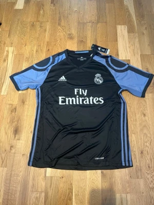 Real Madrid 15/16 away kit X Ronaldo 7 - Snygg svart Real Madrid bortatröja från Adidas med ljusblå detaljer och tryck. Tröjan har klubbmärke, Adidas-logga och Fly Emirates-tryck fram samt Ronaldo 7 på ryggen. Tillverkad i lätt och ventilerande climacool-material, perfekt för fotbollsträning eller match.