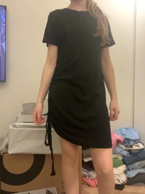 Svart kort klänning med snörning - Svart kortärmad klänning i mjukt tyg med cool snörningsdetalj på sidan som ger en snygg rynkad effekt. Mycket bra skick storlek S. 