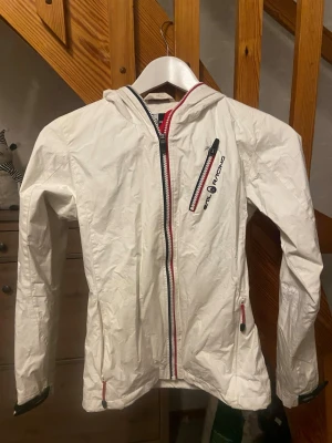 Vit vindjacka Sail Racing med huva - Säljer en vit vindjacka från Sail Racing med huva och coola detaljer. Jackan har logga på bröstet och ärmen samt SR77-brodyr på huvan. Lätt material och perfekt för blåsiga dagar. Snygg passform och långärmad modell. Storlek xs men upplever den mindre. Pris går att diskutera.