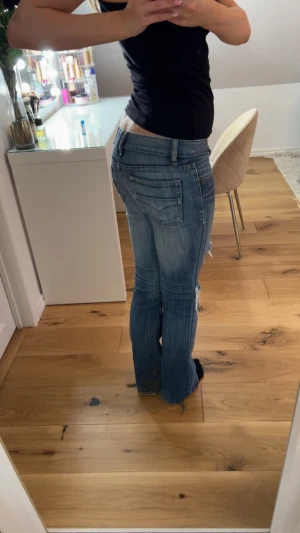 Blå slitna bootcut jeans - Säljer ett par blå bootcut jeans med slitningar och hål på knäna. Jeansen har låg midja, klassiska fem fickor och råa kanter vid benslut. Perfekta för en avslappnad och trendig look. Skriv för fler frågor och bilder! 