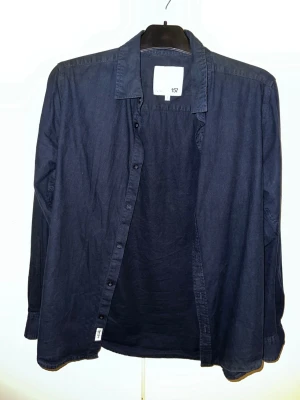 Mörkblå skjorta från 157 - Stilren mörkblå skjorta från 157 med klassisk krage och knäppning framtill. Skjortan är långärmad och har en avslappnad passform, perfekt att bära öppen eller stängd. Tillverkad i mjuk bomull för en bekväm känsla. Behöver bara strykas