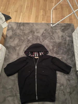 Svart burberry hoodie med rand - En svart hoodie med dragkedja och huva från burberry. Insidan av huvan har ett snyggt randigt foder i vitt, rött och svart. Hoodien har dragsko i huvan och två fickor framtill. Perfekt för en avslappnad och stilren look. riktigt fräsch och ny skick  BYTEN FUNKAR!!!!