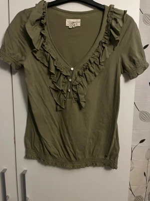 Olivgrön blus med volang och knappar - Supersöt olivgrön blus från Ralph Lauren med volangdetaljer längs v-ringningen och små pärlknappar framtill. Kortärmad modell med resår i ärmslut och nederkant. Perfekt för dig som gillar romantiska detaljer och mjukt material.