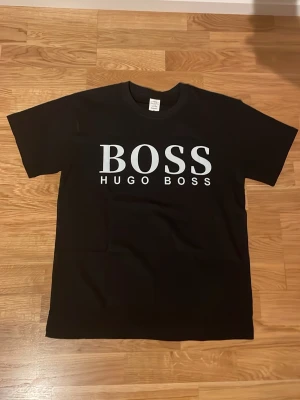 Svart Hugo Boss t-shirt med tryck - Snygg svart t-shirt med stort vitt BOSS-tryck på bröstet. Klassisk rak passform och rund halsringning. Tillverkad i mjuk bomull som känns skön mot huden. Perfekt för en clean och stilren look.