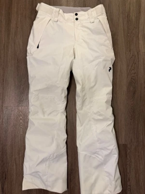 Vita täckbyxor från Peak Performance - Säljer ett par snygga vita täckbyxor från Peak Performance. Byxorna har vida ben, bälteshällor, två framfickor med dragkedja och en liten logga på låret. Perfekta för vinterns alla äventyr i backen eller snön. Materialet är slitstarkt och vattenavvisande.