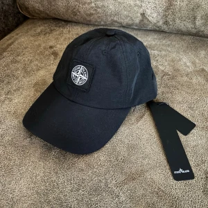 Svart keps från Stone Island - Snygg svart keps från Stone Island med klassisk böjd skärm och ikonisk kompasslogga framtill. Justerbar rem baktill för perfekt passform. Tillverkad i tekniskt tyg som passar till alla streetwear-outfits.