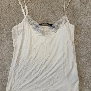 Vit spetsdetaljerad linnetopp - Säljer en vit linnetopp från Black Label Collection med tunna axelband och fina spetsdetaljer längs urringningen. Perfekt att styla lager på lager eller bära som den är. Enkel och stilren design med feminin touch.