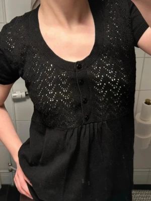 Svart stickad topp från Mingel - Snygg svart stickad topp från Mingel med korta ärmar och rund halsringning. Överdelen har ett fint hålmönster och knappar framtill. Perfekt till jeans eller kjol för en trendig look. Materialet känns mjukt och följsamt.