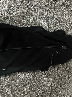 Svart fleeceväst från Peak Performance - Säljer en svart fleeceväst från Peak Performance med hel dragkedja framtill. Västen har en sportig look och är tillverkad i mjukt fleecematerial. Perfekt att slänga över en hoodie eller tröja för extra värme. Diskret logga på bröstet.