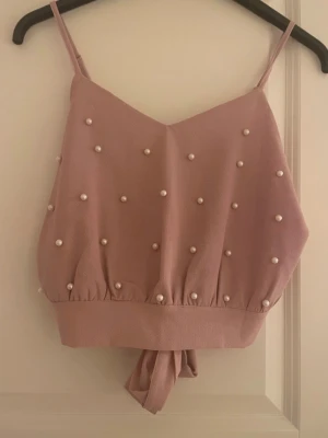Rosa linnetopp med pärlor - Supersöt ljusrosa linnetopp med smala axelband och dekorativa pärlor framtill. Toppen har en croppad passform och knyt i midjan för extra detalj. Perfekt för dig som vill ha en feminin och trendig look.