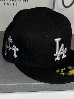 Svart LA keps New Era 59Fifty - Svart fitted keps från New Era 59Fifty med LA-logga i vitt framtill, broderade kors på sidan och vingar samt halo-detalj bak. Kepsen har platt skärm och MLB-logga bak. Materialet är klassisk bomullstwill.
