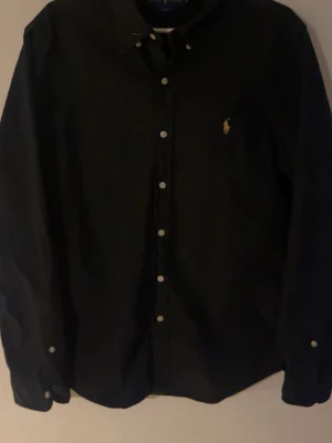 Ralph Lauren skjorta  - Snygg svart skjorta med guldig detalj.     Använt fåtal gånger, inga defekter.                   Priset är inte hugget i sten så det är bara att skriva och fråga!