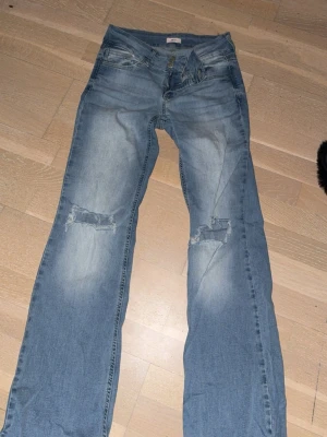 Ljusblå slitna bootcut jeans - Ljusblå bootcut jeans med slitningar över båda knäna och tvättad effekt. Jeansen har klassisk femficksdesign, knapp och dragkedja framtill. Perfekt för dig som gillar en avslappnad och trendig look.