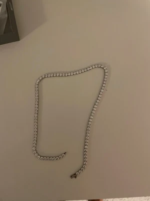 Cernucci halsband med vita stenar - Säljer ett snyggt halsband från Cernucci med vita, gnistrande stenar i hela kedjan. Halsbandet har en klassisk tennisdesign och stängs med ett smidigt lås. Perfekt för dig som vill ha något som sticker ut och glänser.
