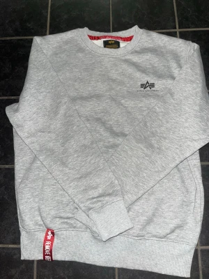Grå sweater från Alpha Industries, Storleken är S - Säljer en stilren grå sweatshirt från Alpha Industries med rund halsringning och diskret logga på bröstet. Tröjan har ribbade muddar vid ärmslut och nederkant samt en röd detalj med texten 'Remove Before Flight' vid nederkanten.