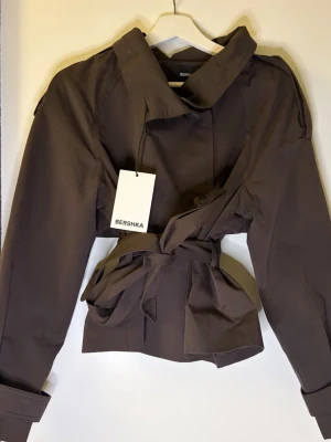 Brun trenchcoat från Bershka - Kort brun trenchcoat från Bershka med bred krage, dubbelknäppning och knytskärp i midjan. Jackan har axelklaffar och justerbara ärmslut för en trendig look. Tillverkad i ett slätt tyg som ger en stilren känsla.