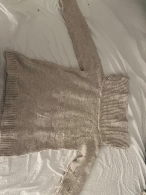 Beige off shoulder stickad tröja - Mysig beige stickad tröja med bred polokrage som kan bäras off shoulder. Tröjan har långa ärmar och ribbstickade muddar vid ärmslut och nederkant. Perfekt för kyliga dagar och ger en avslappnad men trendig look.