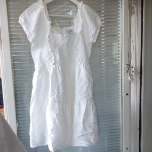 Vit kort klänning med volang - Söt vit klänning från med volangdetaljer och kort ärm. Klänningen har en luftig passform och är tillverkad i bomull, perfekt för varma dagar. Fyrkantig ringning och dragkedja i ryggen ger en stilren look.