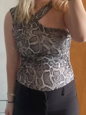 Ormmönstrad asymmetrisk body - Snygg body med ormmönster i grått och svart. Asymmetrisk design med en axel och en bred axelrem som korsar över bröstet. Stretchigt material som sitter tight och framhäver figuren. Perfekt för dig som vill sticka ut lite extra.