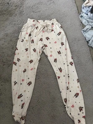 Vita julpyjamasbyxor med mönster - Mysiga vita pyjamasset med gulligt julmönster av små figurer, stjärnor och texten 'Merry Christmas' i rött. Byxorna har resår i midjan med dragsko och muddar vid bensluten. Perfekta för vinterkvällar och julmys!