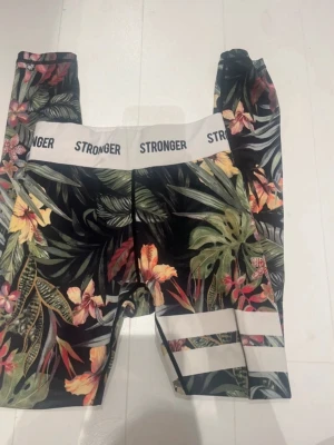 Blommiga tights från Stronger - Snygga träningstights från Stronger med tropiskt blommönster i grönt, gult, rött och orange på svart botten. Bred vit resår i midjan med logga och vita ränder på ena benet. Tillverkade i stretchigt funktionsmaterial som passar perfekt till gymmet eller löpning.