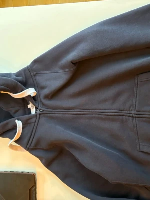 Mörkblå Russedress hoodie med dragkedja - Mörkblå hoodie från Russedress med vit dragsko i huvan och dragkedja framtill. Tryck med Russedress-logga på ena ärmen. Tillverkad i mjukt sweatshirtmaterial och har fickor på sidorna. Perfekt för chill dagar och streetwear-stil.