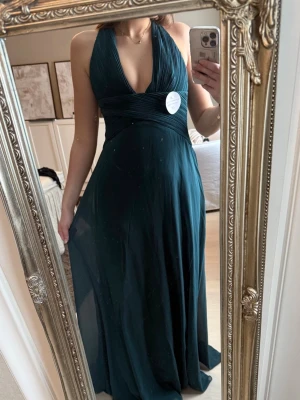 Mörkgrön långklänning  - Elegant mörkgrön långklänning från oh hello clothing, med djup v-ringning och korsade axelband i ryggen. Klänningen har ett flytande fall och är tillverkad i ett mjukt, lätt material som ger en lyxig känsla. Perfekt för speciella tillfällen.