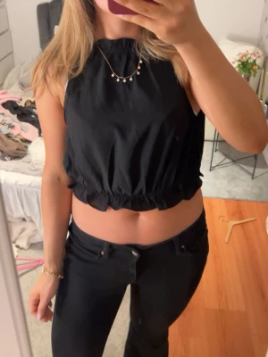 Svart croppad topp med knyt i ryggen - Snygg svart croppad topp med resår i midjan och knytning i ryggen. Toppen är ärmlös och har en avslappnad passform, perfekt för en trendig look. Materialet känns lätt och bekvämt mot huden.