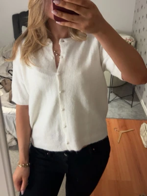 Vit och blå Kortärmad kofta med pärlknappar - Snygg kortärmad kofta i mjukt material, finns i både vitt och mörkblått. Koftan har raka ärmar, rund halsringning och pärlknappar framtill som ger en elegant touch. Perfekt att styla över ett linne eller t-shirt för en trendig look.