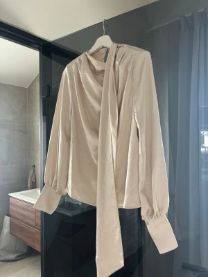 Beige omlottblus med knytband - Stilren beige blus i sidenliknande material med långa puffärmar och breda manschetter. Blusen har ett elegant knytband vid halsen och omlott-detalj framtill. 