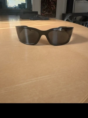 Svarta solglasögon från Prada - Snygga svarta solglasögon från Prada med bred båge och mörka glas. Modellen har en sportig och modern look, perfekt för dig som vill sticka ut. Kommer med originalfodral, putsduk och förpackning från Prada. Två repor på vänster glas som inte påverkar synen. 