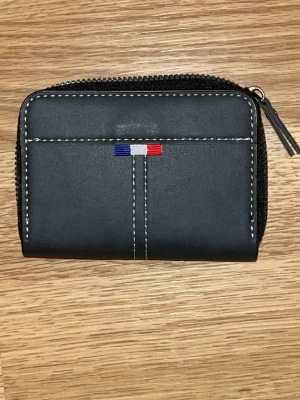 Svart plånbok med dragkedja - Snygg svart plånbok i läderimitation med dragkedja runtom och vita kontrastsömmar. Framsidan har en liten detalj i blått, vitt och rött. Invändigt finns flera fack för kort och sedlar, perfekt för att hålla ordning på allt viktigt.