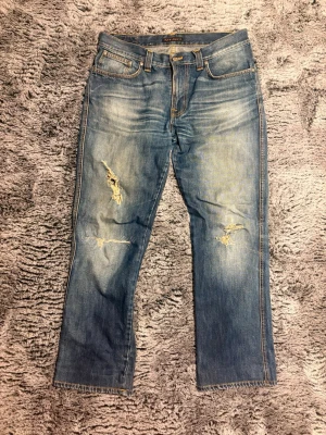 Blå slitna jeans från Nudie Jeans - 103/45 Säljer ett par blå jeans från Nudie Jeans med slitningar och hål på båda benen. Jeansen har klassisk femficksmodell, raka ben och kontrastsömmar. Materialet är denim i bomull och de har en snygg tvättad look med slitna detaljer.