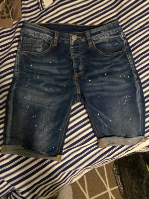 Blå jeansshorts från Kings Will Dream - Säljer ett par blå jeansshorts från Kings Will Dream med coola slitningar och små vita färgstänk. Shortsen har fem fickor, uppvikta benslut och klassisk knappgylf. Perfekta för dig som gillar streetwear och vill ha en avslappnad look.