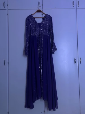 Lång lila glittrig klänning - Säljer en elegant långklänning i lila med glittriga paljetter upptill och långa ärmar. Klänningen har v-ringad hals och ett flödande fall som ger en lyxig känsla. Perfekt för dig som vill sticka ut på festen! Har aldrig använt den har bara provat