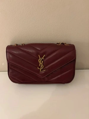 Vinröd YSL handväska i läder - Säljer en vinröd handväska från Yves Saint Laurent i quiltat läder med guldfärgad YSL-logga framtill. Väskan har kedjeremmar i guld och en klassisk, rektangulär form. Perfekt för dig som vill ha en lyxig accessoar med ikonisk design.