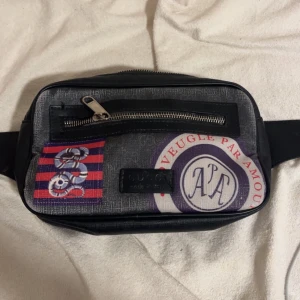 Gucci magväska med tryck och dragkedja - Snygg magväska från Gucci i svart med GG-mönster på baksidan och coolt tryck framtill. Väskan har en silverfärgad dragkedja och justerbart midjeband. Materialet är canvas med detaljer i läder. Perfekt accessoar för att lyfta din outfit.