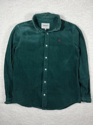 Grön manchester skjorta Carhartt - Mörkgrön manchester skjorta från Carhartt med klassisk krage och vita knappar framtill. Skjortan har lång ärm och en liten röd Carhartt-logga broderad på bröstet. Perfekt för dig som gillar en avslappnad men snygg stil.