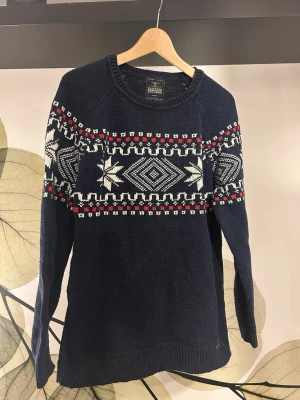 Mörkblå Hampton Republic stickad tröja med mönster - Mysig mörkblå stickad tröja från Hampton Republic med vitt och rött nordiskt mönster över bröstet och ryggen. Tröjan har rund halsringning, långa ärmar och ribbstickade muddar. Perfekt för kalla dagar och ger en klassisk vinterlook.