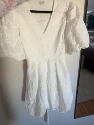 Vit kortklänning med puffärm - Supersöt vit kortklänning från bubbelroom med puffiga kortärmar, v-ringning och diskret blommigt mönster i tyget och knytning i ryggen. Klänningen har en figurnära passform i midjan och ballongformad kjol. Perfekt för sommarens alla tillfällen eller studenten!! Säljer då jag valde en annan studentklänning, helt slutsåld🥰