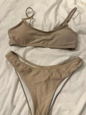 Beige bikini med enkel design - Stilren beige bikini med sportig topp och högt skurna trosor. Båda delarna har tunna axelband och är tillverkade i ett mjukt, stretchigt material som känns bekvämt mot huden. Perfekt för lata dagar på stranden eller poolen.