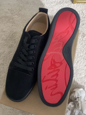 Svarta sneakers från Christian Louboutin - Pris kan sänkas.Snygga svarta sneakers från Christian Louboutin med klassisk röd sula och svart ovandel i mocka. Skorna har svarta skosnören och en stilren, låg profil som passar till många olika outfits. Den röda sulan har den ikoniska Louboutin-signaturen.