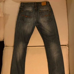 Blå Nudie Jeans med slitningar - Ett par stilrena Nudie Jeans i en klassisk washed blue-tvätt med naturligt ljusare partier som ger ett levande och autentiskt uttryck. Den diskreta slitningen tillför karaktär utan att bli överdriven, vilket gör jeansen enkla att matcha både uppklätt och avslappnat. En tidlös modell med modern känsla och genomarbetade detaljer.  🔷 Washed blue med mjuka, ljusare nyanser för en naturlig denimlook 🔷 Subtila slitningar som ger en balanserad och stilren edge 🔷 Snyggare med tiden 