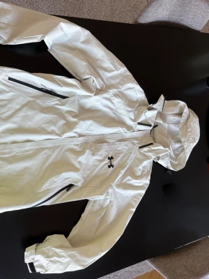 Vit regnjacka från Under Armour - Snygg vit regnjacka från Under Armour med svart logga på bröstet. Jackan har huva, dragkedja framtill och svarta dragkedjor på fickorna. Tillverkad i vattenavvisande material och har justerbara muddar vid ärmsluten. Perfekt för regniga dagar.