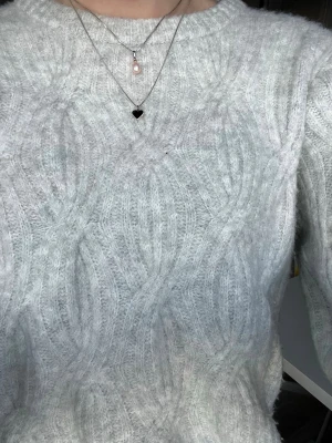 Ljusgrå kabelstickad tröja från Vila - Mysig ljusgrå kabelstickad tröja från Vila med rund halsringning och mjukt, fluffigt material. Tröjan har ett klassiskt stickat mönster och ribbade muddar vid halsen. Perfekt för kyliga dagar och enkel att matcha med olika outfits.