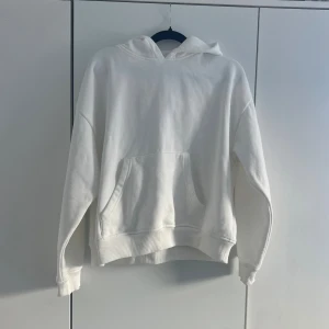 Vit hoodie - Säljer en clean vit hoodie med stor ficka framtill och ribbade muddar vid ärmslut och midja. Hoodien har en klassisk huva utan snören och är tillverkad i mjukt bomullsliknande material. Perfekt för en chill och avslappnad stil. Knappt använd 