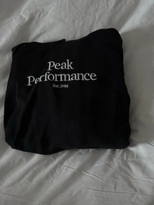 Svart hoodie Peak Performance - Säljer en svart hoodie från Peak Performance med vit broderad logga och texten 'Est. 1986' på bröstet. Klassisk modell med huva och mjukt material, perfekt för en avslappnad stil. Passar dig som gillar stilrena och sportiga plagg.