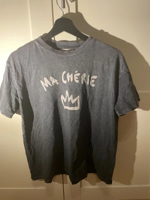 Grå t-shirt med tryck från Gina Tricot - Säljer en grå t-shirt från Gina Tricot med texten 'Ma Chérie' och en krona tryckt på bröstet. T-shirten har korta ärmar, rund halsringning och är gjord i mjuk bomull. Perfekt för en avslappnad och trendig look.