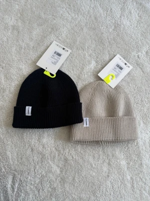 Två ribbstickade mössor från Craft - Två stilrena ribbstickade mössor från Craft, en i svart och en i beige. Båda har uppvikt kant och liten vit logotyp-tag på sidan. Perfekta för kalla dagar och passar till de flesta outfits. Materialet ser ut att vara mjukt och stretchigt, troligen akryl eller ullmix.