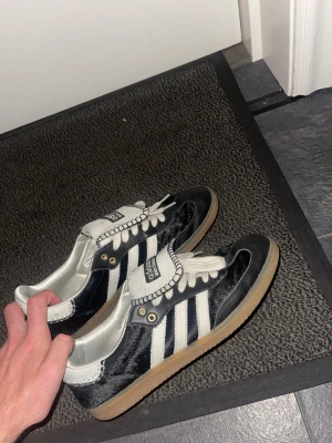 Adidas Samba OG Wales bonner - Säljer ett par klassiska Adidas Samba OG sneakers i svart och vitt med de ikoniska tre ränderna på sidan. Skorna har vit plös med svart Adidas-logga, vita skosnören och gummisula. Perfekta för dig som gillar retrostil och streetwear.